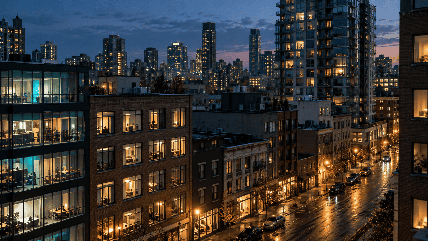 A city block at dusk with lit office windows, storefronts, and apartment buildings